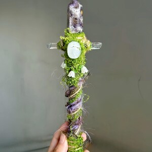 May include: A decorative wand featuring a pointed amethyst crystal at the top, a horizontal clear quartz crossbar, and a handle wrapped in green moss, twine, and purple fibers. The wand is adorned with a central white oval stone and small white flowers.