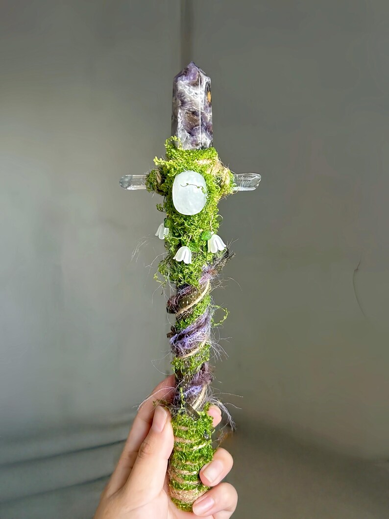 May include: A decorative wand featuring a pointed amethyst crystal at the top, a horizontal clear quartz crossbar, and a handle wrapped in green moss, twine, and purple fibers. The wand is adorned with a central white oval stone and small white flowers.
