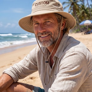 May include: A beige bucket hat featuring the word CUSTOM embroidered in dark brown thread on the front. The hat includes a wide brim and an adjustable chin strap with a black toggle, designed for sun protection and outdoor wear.