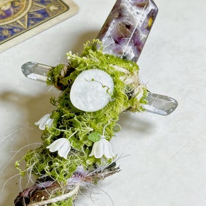 May include: A decorative wand featuring a faceted purple and white crystal point at the top, wrapped in green moss and twine. The handle includes a small white circular stone, three white bell-shaped flower charms, and purple fibers.