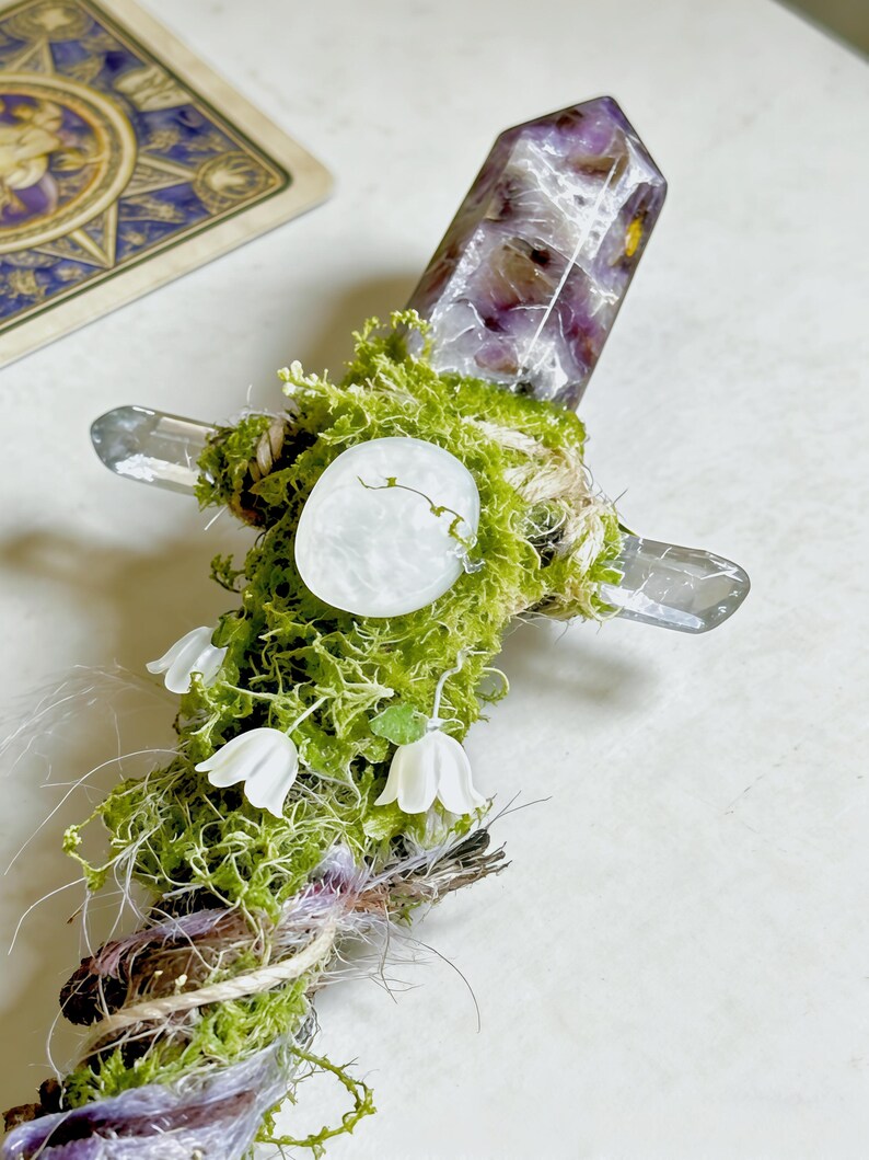 May include: A decorative wand featuring a faceted purple and white crystal point at the top, wrapped in green moss and twine. The handle includes a small white circular stone, three white bell-shaped flower charms, and purple fibers.