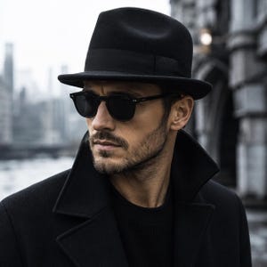Puede incluir: Un sombrero fedora de fieltro negro con una cinta de grosgrain negra a juego, combinado con gafas de sol de cristales oscuros y montura negra. El conjunto incluye un abrigo negro con cuello sobre una camisa negra, creando un aspecto monocromático.