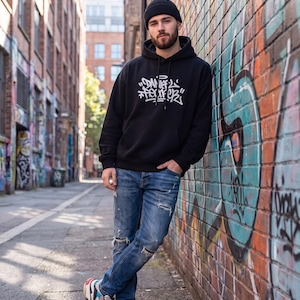 Może przedstawiać: Czarna bluza z kapturem z białym napisem w stylu graffiti na piersi, niebieskie przecierane dżinsy z rozdarciami na kolanach oraz masywne, wielokolorowe sneakersy. Na głowie znajduje się ciemnoszara czapka z dzianiny.