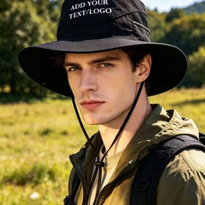 Peut inclure: Un chapeau bob noir doté d'un large bord, de panneaux latéraux en maille et d'une mentonnière réglable avec un cordon de serrage. L'avant du chapeau affiche le texte blanc ADD YOUR TEXT/LOGO. Le chapeau est fait d'un tissu léger et résistant.