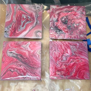 May include: Four square coasters with a pink, white, and gray abstract swirl design. The coasters are made with a resin-like material and have a glossy finish.
