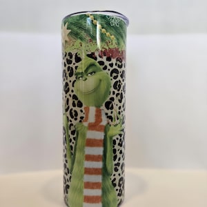 May include: A stainless steel tumbler featuring a green Grinch character with a striped scarf, set against a leopard print background. The top of the tumbler has a Christmas tree design. The tumbler is designed to hold approximately 20 ounces.