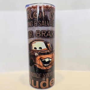 Pode incluir: Um copo de a&ccedil;o inoxid&aacute;vel com uma imagem de desenho animado do Mate do filme Carros. O copo tem o texto "Mater B Towing & Salvage" e "Made Your Day Dude" em letras marrons.