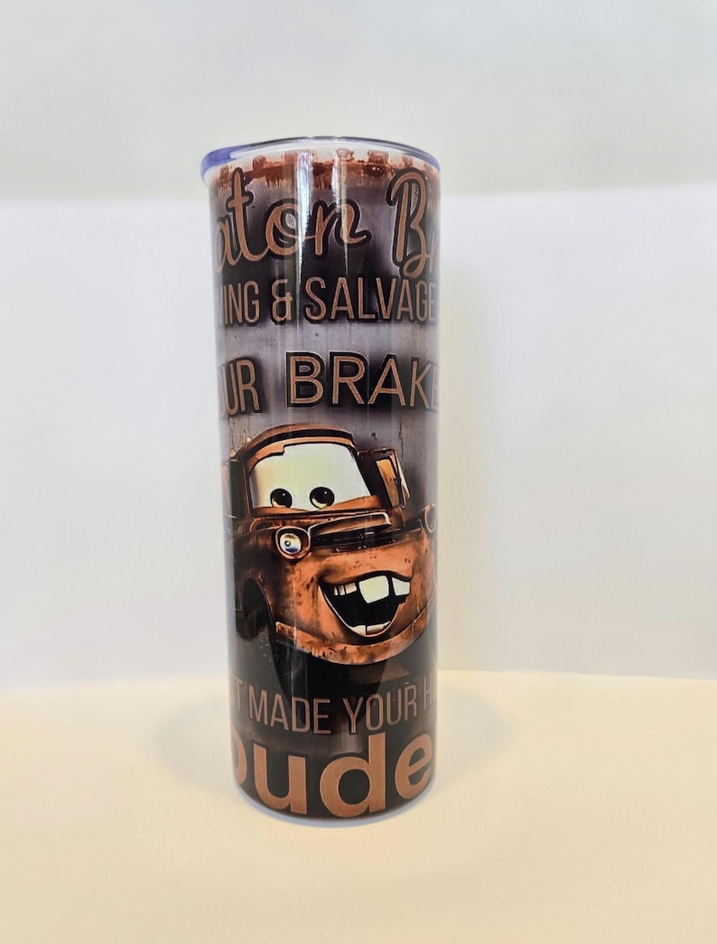 Pode incluir: Um copo de a&ccedil;o inoxid&aacute;vel com uma imagem de desenho animado do Mate do filme Carros. O copo tem o texto "Mater B Towing & Salvage" e "Made Your Day Dude" em letras marrons.