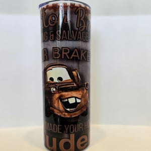 Pode incluir: Um copo cil&iacute;ndrico com uma imagem de desenho animado de Mate do filme Carros. O copo tem o texto "Mater's Towing & Salvage" e "Made Your Home Rude". O copo &eacute; castanho e tem uma tampa prateada.