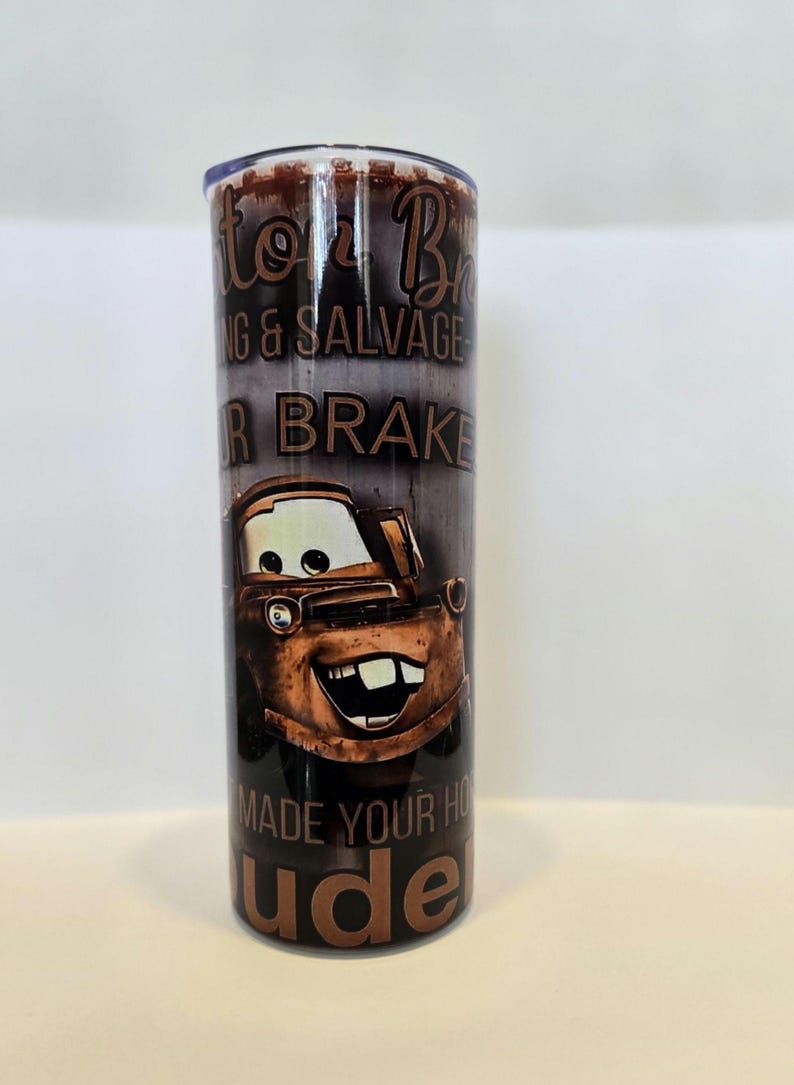 Pode incluir: Um copo cil&iacute;ndrico com uma imagem de desenho animado de Mate do filme Carros. O copo tem o texto "Mater's Towing & Salvage" e "Made Your Home Rude". O copo &eacute; castanho e tem uma tampa prateada.