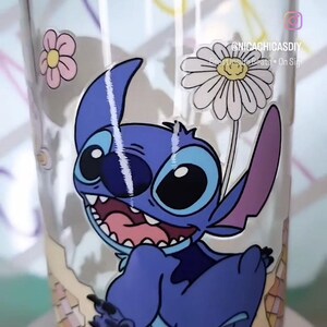 May include: A clear glass jar featuring a blue and white cartoon character of Stitch from Lilo & Stitch. The character is peeking out of a broken, colourful mosaic design. The jar has a wooden lid and a white daisy with a yellow centre.