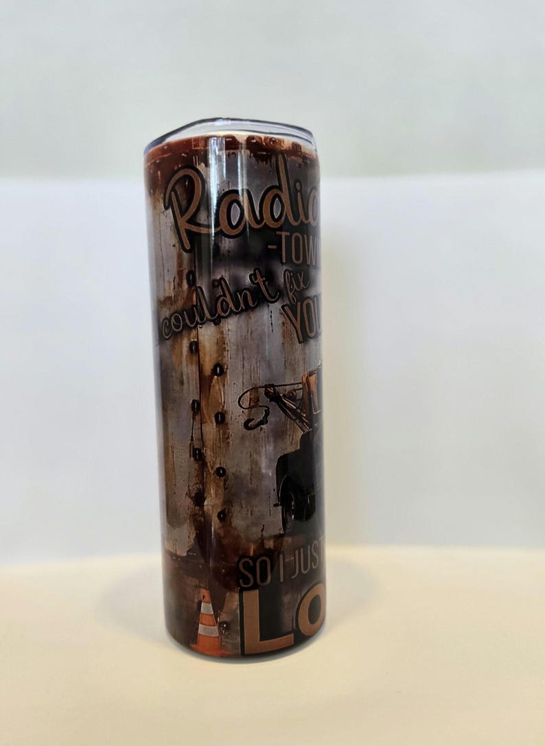 Pode incluir: Caneca de a&ccedil;o inoxid&aacute;vel com um design de metal desgastado. A caneca apresenta o texto "Radio Tow Couldn't Fix You, So I Just Love You" em marrom e branco. Um gr&aacute;fico de reboque e um cone de tr&acirc;nsito laranja tamb&eacute;m s&atilde;o vis&iacute;veis.