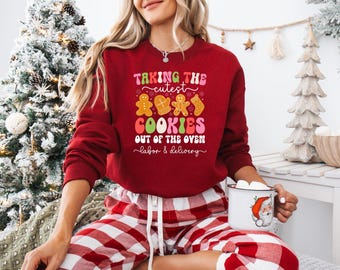 May include: A red sweatshirt with the text "Taking the cutest cookies out of the oven labor & delivery" in a colorful design. The sweatshirt features gingerbread men, mittens, and a stocking graphic. The person is wearing red and white plaid pajama pants.