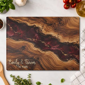 May include: A rectangular wooden cutting board featuring a prominent, dark red resin inlay with gold flecks running diagonally across the centre. The board is engraved with the text Emily & Trevor and 40 YEARS in a light, script and sans-serif font.