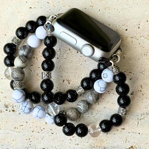 May include: Silver Apple Watch with a black screen, attached to a beaded bracelet. The bracelet features black, white, and gray beads, with decorative silver accents. The watch band is designed to wrap around the wrist.