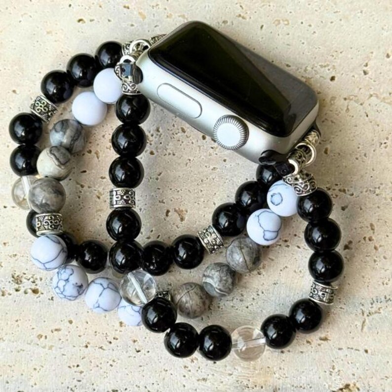 May include: Silver Apple Watch with a black screen, attached to a beaded bracelet. The bracelet features black, white, and gray beads, with decorative silver accents. The watch band is designed to wrap around the wrist.
