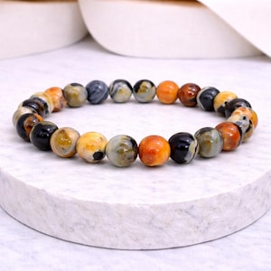 Könnte beinhalten: Ein Armband mit runden Perlen in verschiedenen Farben. Die Perlen sind in Orange, Schwarz, Gelb und Grau gehalten und bilden ein einzigartiges Design. Das Armband liegt auf einer weißen Oberfläche.