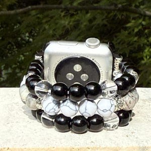 May include: A silver Apple Watch with a black band is surrounded by three beaded bracelets. The bracelets feature black, white, and gray beads. The background is a blurred green.