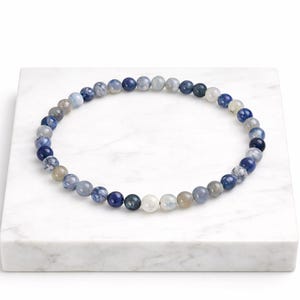May include: A beaded bracelet featuring a variety of blue, gray, and white round beads. The bracelet is displayed on a white marble surface. The beads vary in color and pattern.