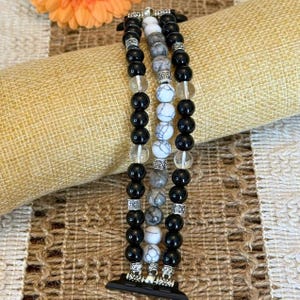May include: A beaded watch band featuring three strands of black, white, and clear beads. The band has silver-colored connectors and is designed to fit a smartwatch. The beads are round and vary in size.