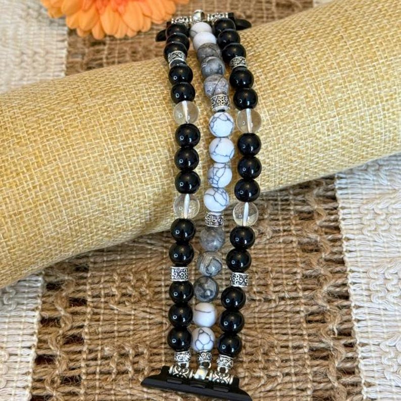 May include: A beaded watch band featuring three strands of black, white, and clear beads. The band has silver-colored connectors and is designed to fit a smartwatch. The beads are round and vary in size.