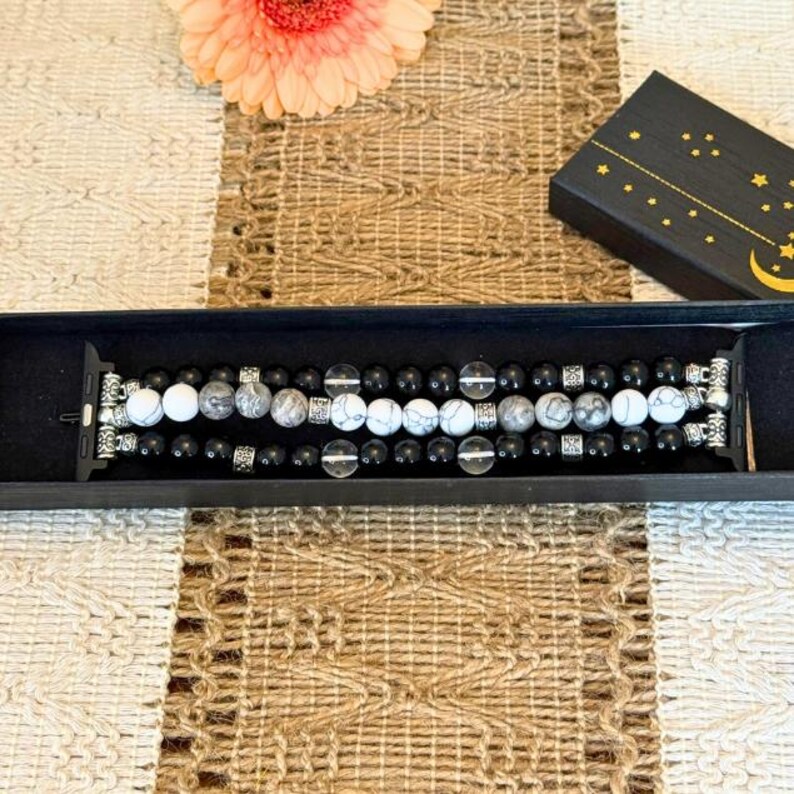 May include: A beaded Apple Watch band featuring black, white, and gray stones. The band is displayed in a black box with a floral accent and a decorative box with a celestial design.