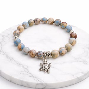 Puede incluir: Una pulsera de cuentas redondas de jaspe multicolor en tonos azules, marrones y beige. Un dije de tortuga plateado cuelga de la pulsera. La pulsera se exhibe sobre una superficie de mármol blanco.