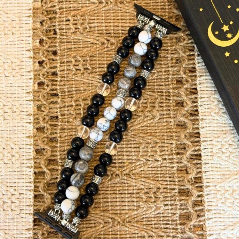 May include: A beaded Apple Watch band featuring black, white, and gray stone beads. The band has a decorative silver-colored metal clasp and connectors. The beads are strung together with clear, round beads. The band is displayed on a textured, neutral-toned surface.