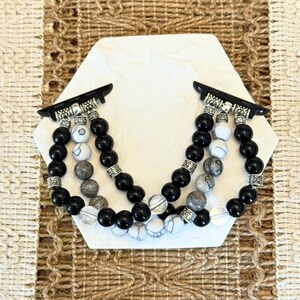 May include: Black and white beaded Apple Watch band. The band features black, white, and gray beads with silver-colored metal accents. The band is displayed on a white hexagonal surface.