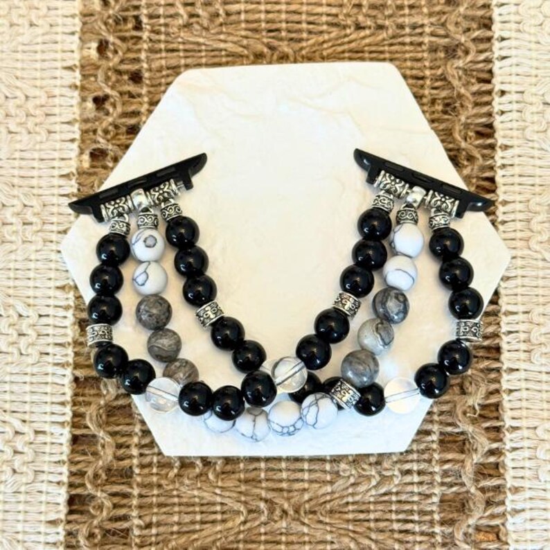 May include: Black and white beaded Apple Watch band. The band features black, white, and gray beads with silver-colored metal accents. The band is displayed on a white hexagonal surface.