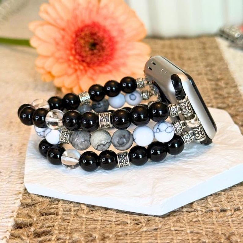 May include: Stack of three beaded bracelets with an Apple Watch attached. The bracelets feature black, white, and gray beads with silver accents. A peach gerbera daisy is in the background.
