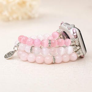 May include: A beaded Apple Watch band featuring three strands of pink and white beads. The band has silver-colored accents and a small charm. The watch itself is silver with pink details.