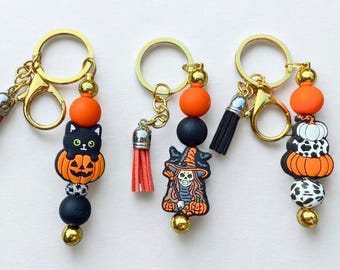 Puede incluir: Llaveros temáticos de Halloween con herrajes dorados. Cada llavero presenta un dije único: un gato negro en una calabaza, una bruja y calabazas apiladas con estampado de vaca. Cuentas y borlas naranjas, negras y blancas añaden detalles.