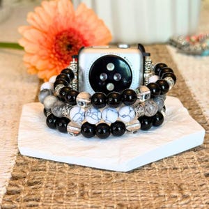 May include: Stack of three beaded bracelets with black, white, and gray stones. The bracelets are displayed with a silver smartwatch and a vibrant orange flower. The bracelets are on a white geometric stand.