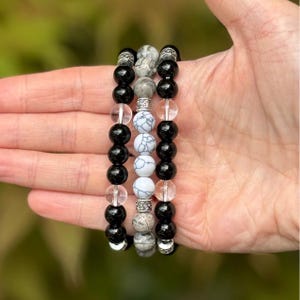 May include: Three beaded bracelets with black, clear, and white patterned beads. The bracelets are held in a hand, showcasing the different textures and colors of the beads. The bracelets have silver-colored spacers.