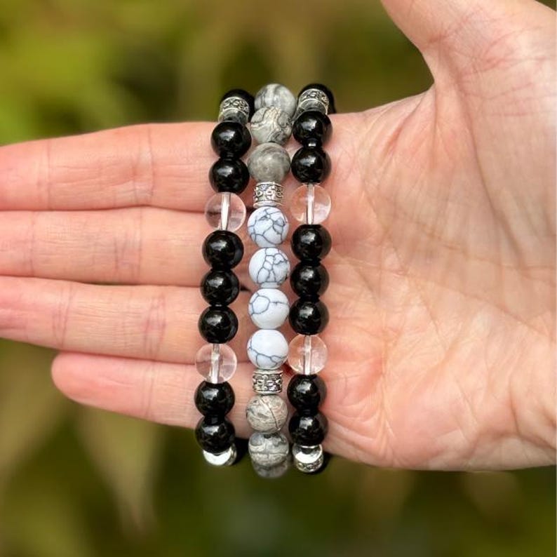 May include: Three beaded bracelets with black, clear, and white patterned beads. The bracelets are held in a hand, showcasing the different textures and colors of the beads. The bracelets have silver-colored spacers.