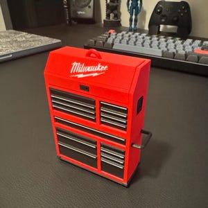 Può includere: Una cassetta degli attrezzi rossa Milwaukee con una maniglia nera e diversi cassetti. La cassetta degli attrezzi è in plastica e ha un logo bianco sul davanti.