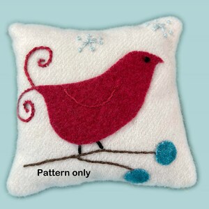 May include: A white felt pillow with a red bird embroidered on it. The bird is perched on a brown branch with blue berries. The background is white with snowflakes embroidered in blue.