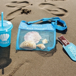 May include: A blue mesh bag with the text "Nicole's Beach Treasures" and a starfish design. The bag is filled with seashells. There is also a blue cup with the name "Nicole" and a blue and white seashell design.