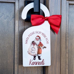 Op de afbeelding: Witte deurhanger met een rode strik, met een vintage illustratie van de kerstman die een zak met cadeaus vasthoudt. De tekst luidt "Santa Please Stop Here For Kennedi".