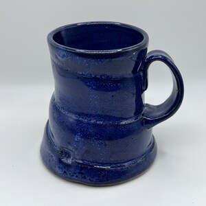 May include: A blue ceramic mug with a unique, slightly asymmetrical shape. The mug has a wide, rounded base and a slightly flared rim. The handle is attached to the side of the mug and curves upwards.