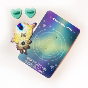 Pu&ograve; includere: Uno spillo in smalto olografico con un astronauta dei cartoni animati con uno schema di colori arcobaleno. Lo spillo &egrave; su una carta di supporto con un design a galassia e il testo "Enamel Pin" e "www.ettoarts.com".