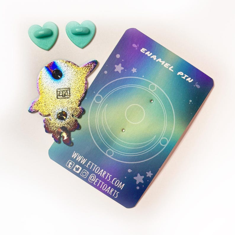 Pu&ograve; includere: Uno spillo in smalto olografico con un astronauta dei cartoni animati con uno schema di colori arcobaleno. Lo spillo &egrave; su una carta di supporto con un design a galassia e il testo "Enamel Pin" e "www.ettoarts.com".