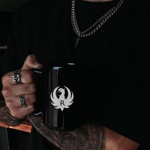 Black Ruger logo mug featuring a white phoenix emblem with letter R. Ideal gun enthusiast mug, Ruger coffee mug, firearms brand drinkware, patriotic mug, military supporter mug, veteran coffee cup, tactical mug, range day mug, unisex coffee mug, office mug, collector mug, American pride mug, shooting sports mug, gun owner gift, second amendment mug, black matte mug, logo coffee cup, everyday use mug.