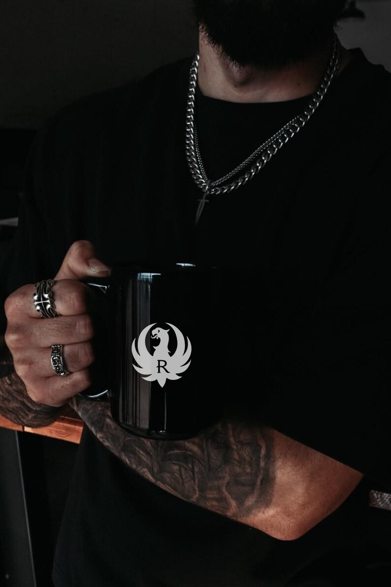 Black Ruger logo mug featuring a white phoenix emblem with letter R. Ideal gun enthusiast mug, Ruger coffee mug, firearms brand drinkware, patriotic mug, military supporter mug, veteran coffee cup, tactical mug, range day mug, unisex coffee mug, office mug, collector mug, American pride mug, shooting sports mug, gun owner gift, second amendment mug, black matte mug, logo coffee cup, everyday use mug.