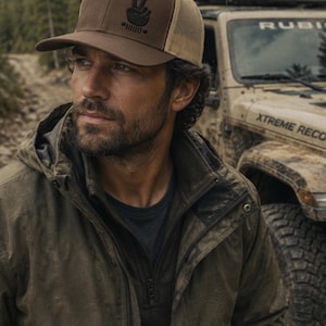 Könnte beinhalten: Braune und beige Trucker-Cap mit schwarzem Peace-Zeichen-Design. Die Kappe hat einen gebogenen Schirm und eine Mesh-Rückseite. Die Person im Bild trägt eine grüne Jacke und steht in der Nähe eines Jeeps.