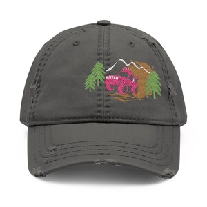 Puede incluir: Gorra de béisbol gris desgastada con un diseño bordado de un vehículo todoterreno rosa, árboles y montañas. La gorra tiene un aspecto desgastado con bordes deshilachados, ofreciendo una estética vintage. Un accesorio elegante para los entusiastas del aire libre.