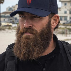 May include: Navy blue baseball cap with a red bull graphic. The cap has a mesh back and a curved brim. The person in the image is wearing a black jacket and has a long beard.