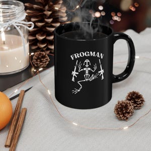 May include: Black ceramic coffee mug with a white frogman skeleton holding two guns and the text "FROGMEN" printed on the front.