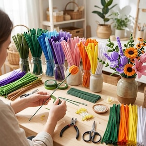 May include: A craft table featuring numerous bundles of colorful pipe cleaners in glass jars, including green, blue, purple, pink, orange, yellow, and white. A bouquet of felt flowers sits in a ceramic vase, alongside floral wire, tape, and tools.